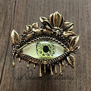 以下が含まれることがあります： アンティーク調の、詳細な目の形をした装飾品。目は、黒い瞳孔を持つ緑色の虹彩を持ち、花と葉のアクセントが付いた金色の金属フレームで囲まれています。フレームの下部には滴るような効果があります。