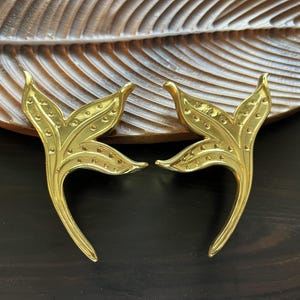 Może przedstawiać: Dwa złote, dekoracyjne przedmioty w kształcie liści. Każdy ma zakrzywioną łodygę i szczegółowy wzór liścia z małymi, równomiernie rozmieszczonymi kropkami. Obiekty umieszczone są na ciemnej drewnianej powierzchni z rzeźbionym wzorem liścia.