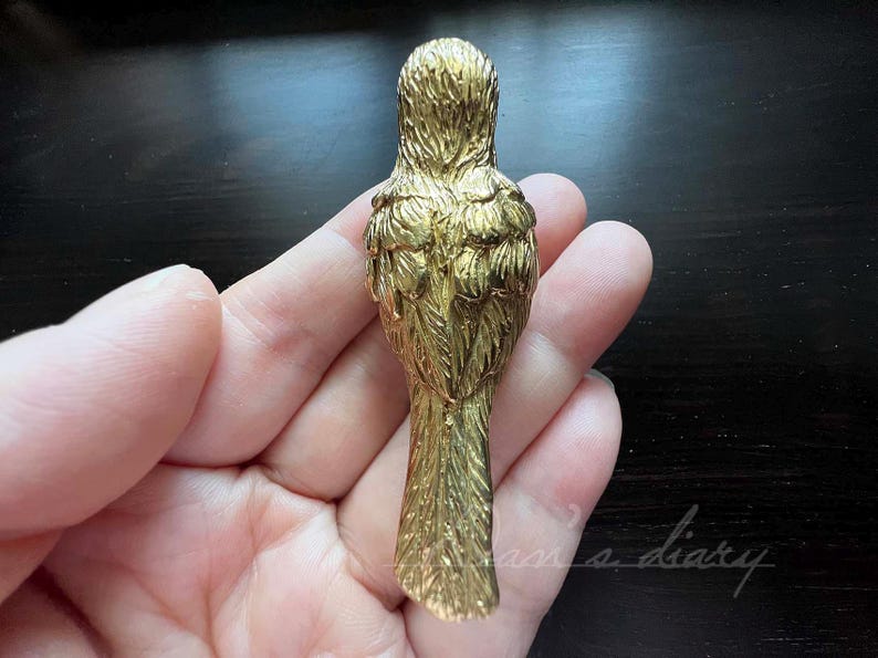 Puede incluir: Una figurita de p&aacute;jaro de metal de color dorado con plumas detalladas. El p&aacute;jaro est&aacute; de espaldas al espectador, con las alas plegadas y las plumas de la cola extendidas.