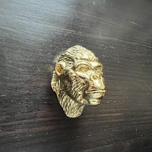 May include: A gold-colored gorilla head sculpture with a detailed face and fur. The sculpture is mounted on a dark wooden surface.
