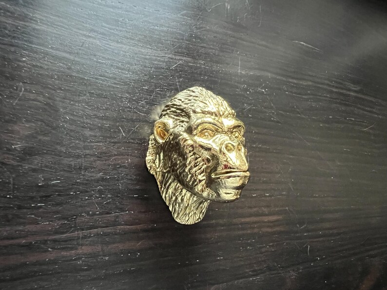 May include: A gold-colored gorilla head sculpture with a detailed face and fur. The sculpture is mounted on a dark wooden surface.