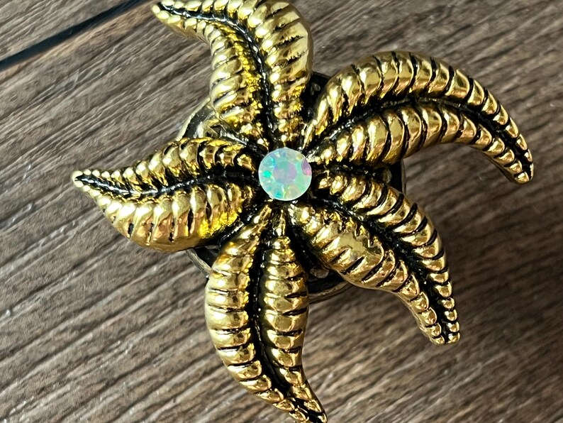 Op de afbeelding: Een goudkleurig, stervormig decoratief object met een centrale iriserende edelsteen. De zeester heeft gedetailleerde, getextureerde segmenten en is geplaatst tegen een houten achtergrond. Het object is waarschijnlijk een decoratieve speld of versiering.