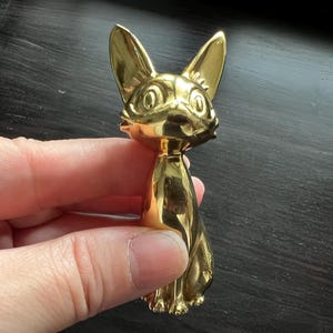 以下が含まれることがあります： 大きな耳と詳細な顔を持つ金色の猫の置物。 猫は座っており、滑らかで反射する表面を持っています。 置物は暗い背景に対して手で持たれています。