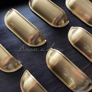 Może przedstawiać: Zestaw ośmiu złotych uchwytów do szuflad szafek o zaokrąglonym, owalnym kształcie. Uchwyty są wykonane z metalu i mają gładkie, polerowane wykończenie.