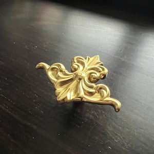 Może przedstawiać: Złoty metalowy akcent dekoracyjny z kwiatowym wzorem. Akcent ma szczegółowy, ozdobny wzór z centralnym kwiatem i zwijającymi się liśćmi.