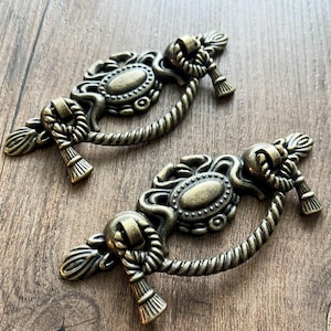 May include: Two antique bronze drawer pulls with a rope design. Each pull features an oval centre and decorative accents. The pulls are set against a wooden background, showcasing their vintage aesthetic.