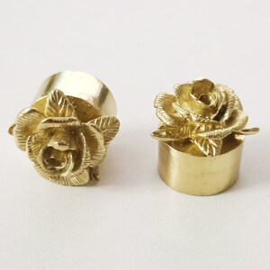 May include: Two gold-colored metal bottle stoppers with a rose design. The stoppers are round and have a smooth surface. The roses are detailed and have a realistic appearance.