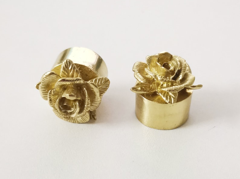 May include: Two gold-colored metal bottle stoppers with a rose design. The stoppers are round and have a smooth surface. The roses are detailed and have a realistic appearance.