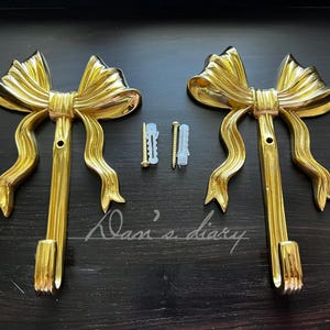May include: Two gold-tone metal hooks shaped like bows. Each hook has a decorative bow with a loop at the top and a hook at the bottom. The hooks are mounted on a dark surface.
