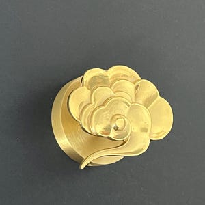 May include: A gold-coloured, cloud-shaped cabinet knob. The knob has a detailed, layered design with a swirling element. The hardware is mounted on a circular base, set against a dark background.