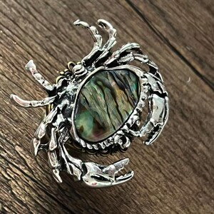 K&ouml;nnte beinhalten: Silberfarbene Brosche in Krabbenform mit einem ovalen Abalone-Muschel-Mittelteil. Die Krallen und der K&ouml;rper der Krabbe sind mit aufwendigen Texturen versehen. Die Abalone-Muschel zeigt schillernde gr&uuml;ne, blaue und braune Farbt&ouml;ne.