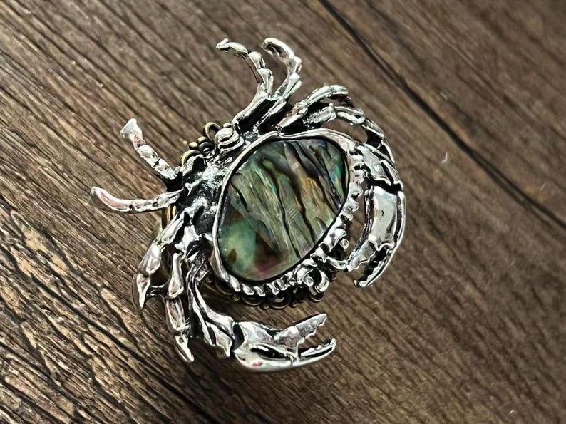 K&ouml;nnte beinhalten: Silberfarbene Brosche in Krabbenform mit einem ovalen Abalone-Muschel-Mittelteil. Die Krallen und der K&ouml;rper der Krabbe sind mit aufwendigen Texturen versehen. Die Abalone-Muschel zeigt schillernde gr&uuml;ne, blaue und braune Farbt&ouml;ne.