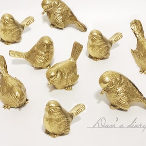 Peut inclure: Une collection de huit figurines d'oiseaux dorées. Les oiseaux sont tous dans des poses différentes, certains sont perchés, d'autres volent et d'autres sont debout. Les oiseaux sont tous faits d'un matériau lisse et brillant.