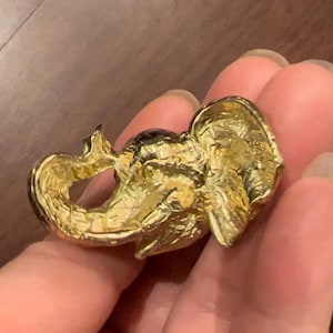 May include: A gold-coloured elephant brooch. The brooch depicts a detailed design of an elephant's head and trunk, with textured surfaces. The elephant's trunk is curled, and the brooch has a pin closure on the back.