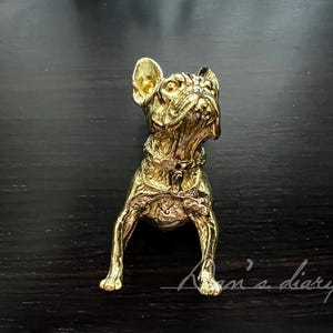 May include: A gold-colored metal figurine of a French Bulldog sitting with its head tilted up. The figurine is detailed with a textured surface and a collar.
