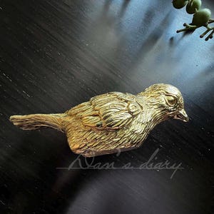 Puede incluir: Una figurita de p&aacute;jaro de metal de color dorado con plumas detalladas. El p&aacute;jaro est&aacute; posado sobre una superficie oscura.