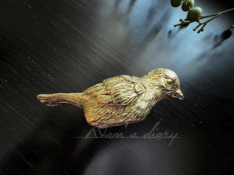 Puede incluir: Una figurita de p&aacute;jaro de metal de color dorado con plumas detalladas. El p&aacute;jaro est&aacute; posado sobre una superficie oscura.