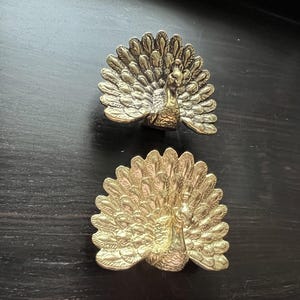 Może przedstawiać: Dwie złote figurki pawia z rozłożonymi ogonami. Pawie są wykonane z metalu i mają szczegółowy wzór.