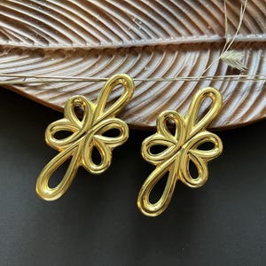 Peut inclure: Une paire de boucles d'oreilles dorées avec un design unique en forme de boucle. Les boucles d'oreilles ont une forme florale avec de multiples boucles et courbes. Elles sont présentées sur un plateau en bois en forme de feuille sur fond sombre.