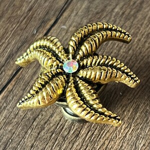 Op de afbeelding: Een goudkleurige decoratieve knop in de vorm van een zeester met een centrale iriserende edelsteen. De knop heeft gedetailleerde ribbels en is waarschijnlijk van metaal. Het ontwerp doet denken aan kustdecoratie.