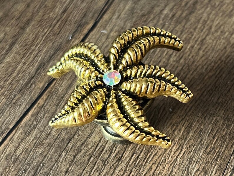 Op de afbeelding: Een goudkleurige decoratieve knop in de vorm van een zeester met een centrale iriserende edelsteen. De knop heeft gedetailleerde ribbels en is waarschijnlijk van metaal. Het ontwerp doet denken aan kustdecoratie.