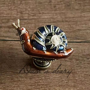 May include: A decorative snail-shaped cabinet knob. The knob features a brown body, a blue and white shell, and gold-colored antennae. The snail is mounted on a decorative brass base, adding a touch of whimsy to any furniture piece.