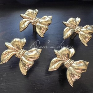 May include: Four gold-colored bow-shaped cabinet knobs. Each knob features a detailed bow design with a textured center. The knobs are arranged on a dark surface, creating a contrast that highlights their metallic finish.