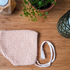 May include: A pink knitted baby bonnet with a white crocheted tie. The bonnet is lying on a wooden surface.