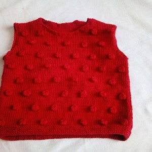 Peut inclure: Un gilet rouge vif en tricot avec un motif en point de fantaisie. Le vêtement sans manches a une encolure arrondie et un ourlet côtelé. Le gilet est probablement pour un enfant ou un bébé.
