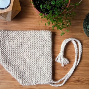 May include: A beige knitted hat with a long white string tie. The hat has a pointed tip and a tassel on the end of the tie.