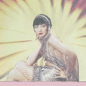 May include: Vintage postcard featuring a seated woman in a beaded gown and headpiece, set against a yellow and red fan-shaped background. The woman is posed with her hand near her face, with a red nail polish. The postcard has a distressed, aged appearance.
