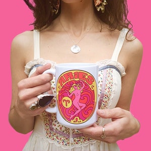 May include: White ceramic mug with a colorful illustration of an Aries zodiac sign and the word "Aries" in a retro font. The illustration features a woman with long blonde hair wearing a pink dress and a yellow crown.