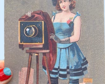 Könnte beinhalten: Eine rechteckige Karte im Vintage-Stil mit einer farbigen Illustration einer Person, die neben einer großen, dunklen Kastenkamera auf einem Holzstativ steht. Die Person trägt ein blaues Kleid mit schwarzen Streifen, dunkle Leggings und ein blaues Barett. Das Wort Photographer ist unten aufgedruckt.