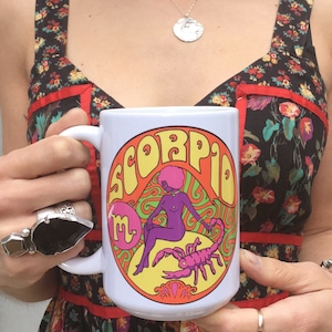 May include: White ceramic mug with a colorful graphic of a woman with a scorpion and the word "Scorpio" in a psychedelic font.