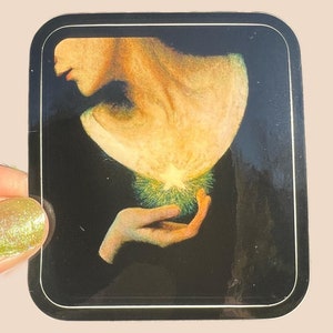 May include: A sticker featuring a woman's hand holding a glowing orb of light. The orb is green and yellow with a white center. The woman's hand is pale and the background is black.