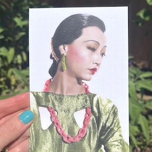 May include: A postcard showing a vintage photograph of a woman with dark hair, red lipstick, and blush. She wears a green dress and a pink necklace. The image is held by a hand with a blue painted nail.