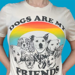 May include: Cream-colored t-shirt with the text "DOGS ARE MY FRIENDS" in black, arched above a rainbow graphic. The graphic features a collection of dog illustrations in black and white.