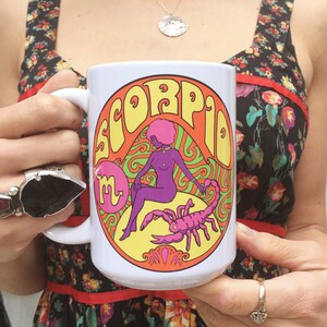 May include: White ceramic mug with a colorful graphic of a woman and a scorpion. The graphic includes the word "Scorpio" in a psychedelic font.