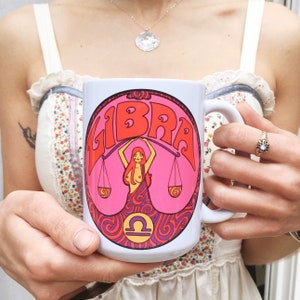 May include: White ceramic mug with a pink and purple design featuring a woman holding a scale and the word "LIBRA" in a retro font.