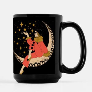 May include: A black ceramic mug with a white crescent moon and a woman in a red and yellow costume reaching for stars. The mug has a black handle.