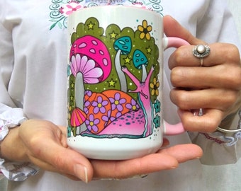 May include: White ceramic mug with a pink, green, and blue illustration of a snail and mushrooms. The mug has a pink handle.
