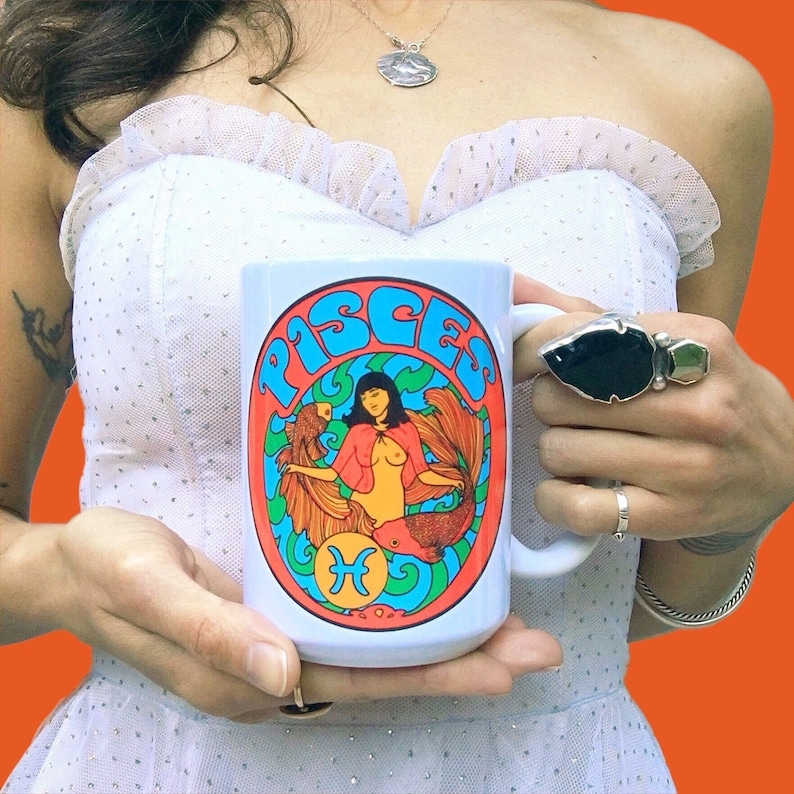 May include: White ceramic mug with a colorful illustration of a mermaid with the word "Pisces" in a retro font. The mermaid is surrounded by fish and seaweed.