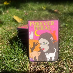 May include: A pink and black sticker with the text "FOREVER FERAL" in yellow letters. The sticker features a woman with long black hair and a cat-like face, surrounded by cats.