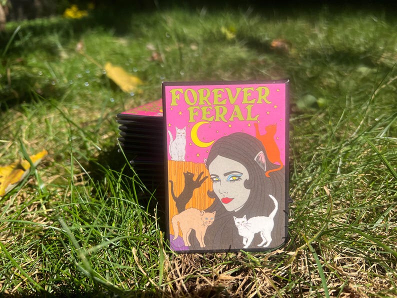 May include: A pink and black sticker with the text "FOREVER FERAL" in yellow letters. The sticker features a woman with long black hair and a cat-like face, surrounded by cats.