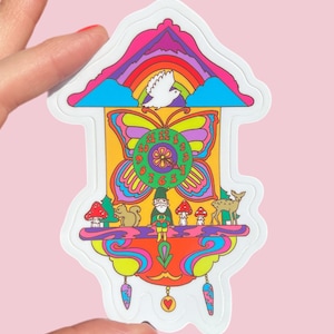Può includere: Un adesivo colorato a forma di orologio a cucù. L'orologio presenta un quadrante a farfalla, un arcobaleno e una colomba bianca. Sono raffigurati anche uno gnomo, uno scoiattolo, un cervo e dei funghi. L'adesivo è tenuto da una persona.