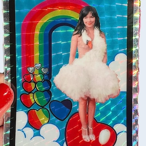 May include: A holographic sticker featuring a woman in a swan dress, standing on a red heart. A rainbow arches above her, with colorful hearts and clouds. The sticker has a black border with a silver, reflective edge.