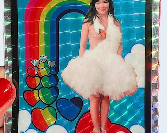 May include: A holographic sticker featuring a woman in a swan dress, standing on a red heart. A rainbow arches above her, with colorful hearts and clouds. The sticker has a black border with a silver, reflective edge.