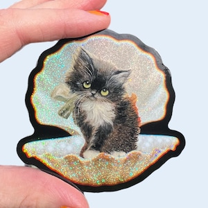 May include: A sticker featuring a fluffy black and white kitten with green eyes, holding a small object. The kitten is set inside a glittery seashell design with a black border. The background is a light blue.
