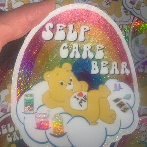 May include: A rainbow glitter sticker featuring a Care Bear with the text "SELF CARE BEAR" and "I &hearts; ME" on its belly. The bear is lying on a cloud with a rainbow background.