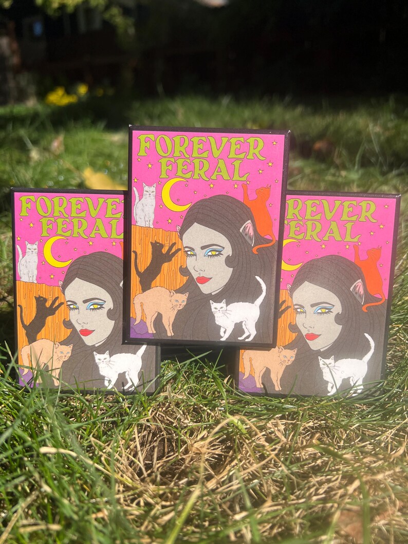May include: Three pink and black square cards with the text "Forever Feral" in yellow letters. Each card features a woman with cat ears and a variety of cats in different colors.